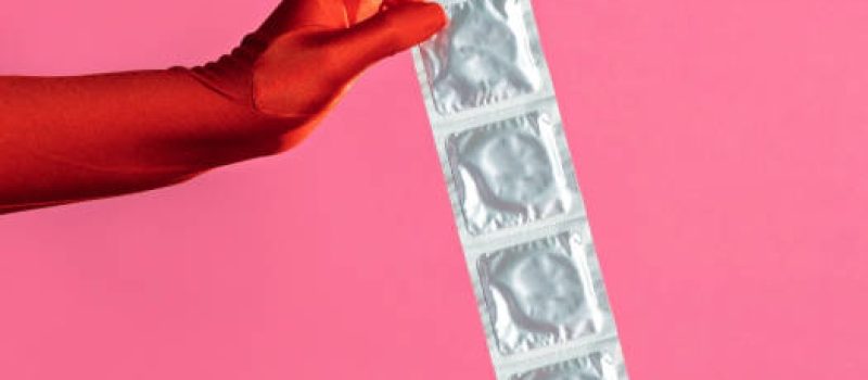 A woman's hand in a red glove holds a pack of condoms on a pink background